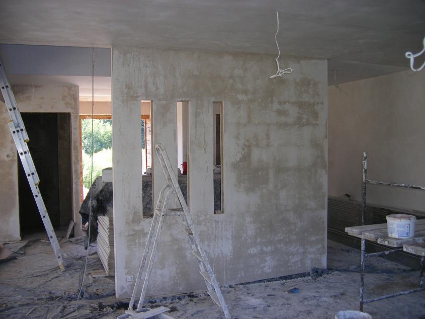 Wnętrze w stanie surowym po nałożeniu tynku, widoczne ślady po tynkowaniu maszynowym, aluminiowa drabina oparta o ścianę z trzema pionowymi otworami, kabel zwisający z sufitu.